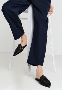 Les mules tressées noires présentent un bout pointu et une semelle plate, avec un design texturé. Associées à un pantalon large bleu marine sur un tabouret.