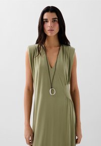 Abito senza maniche verde oliva con scollatura a V, dettaglio arricciato sul lato e una texture liscia. Presenta una lunga collana in pelle marrone con un pendente in argento.