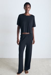 Žena stoji bosa na tepihu, obučena u labavi crni crop top i široke hlače, protiv običnog bijelog zida.