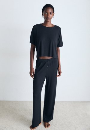 Kvinna som står barfota på heltäckningsmatta och bär en lös svart crop top och vida byxor mot en enkel vit vägg.