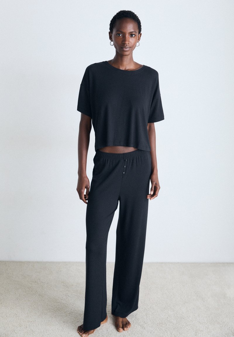 Žena stoji bosa na tepihu, obučena u labavi crni crop top i široke hlače, protiv običnog bijelog zida.