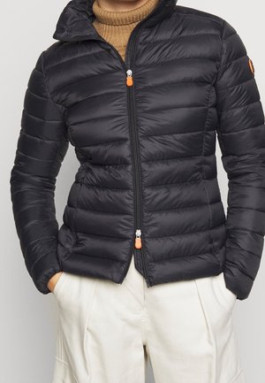 Woman wearing black quilted jacket with orange zipper pulls over brown turtleneck and cream pants, hands in pockets, standing against plain background.