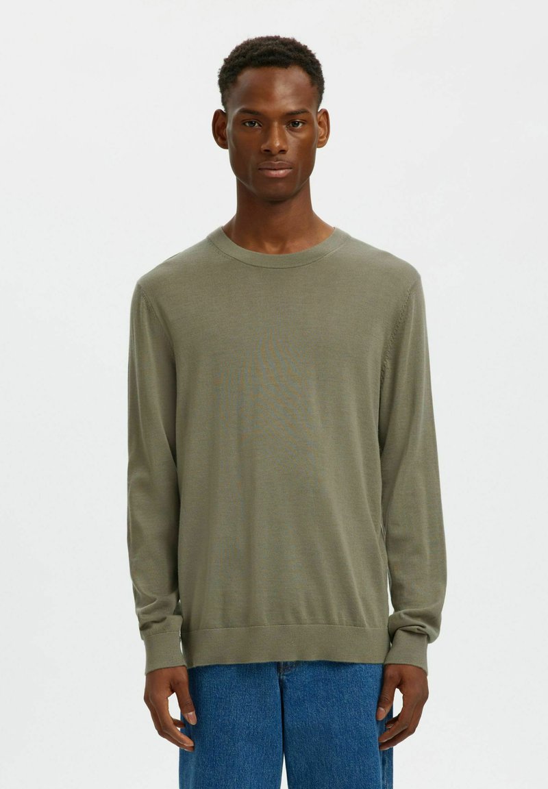 Pull en maille vert olive à manches longues avec un col rond, des poignets et un ourlet côtelés, porté par-dessus un pantalon en denim bleu décontracté.