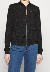 Svart bomberjacka med dragkedja fram, prickigt mönster, elastiska ärmslut och nederkant. Har en rund krage och en diskret logotypaccenter på bröstet.