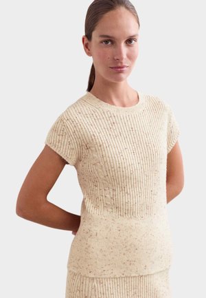 Woman with tied-back hair wearing a short-sleeve beige cable-knit sweater standing with hands behind her back.