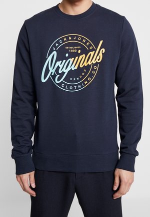 Person iført en marineblå sweatshirt med flerfarvet tekst "Jack & Jones Originals Clothing Co Established 1990 CP NHGN" på brystet.