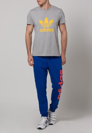 Šedé bavlněné tričko s žlutým logem Adidas, v kombinaci s modrými tepláky s červeným brandingem a bílými sportovními botami se sponami.