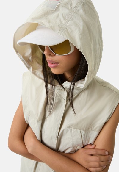 Beige ärmellose Kapuzenjacke mit einem frontalen Knopfverschluss, zwei Brusttaschen und einer weichen Textur; kombiniert mit einer weißen Mütze und großen Sonnenbrillen.