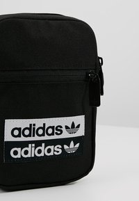 Černý látkový batoh s texturovaným povrchem, který má tři bílé nášivky loga Adidas. Zapínání na zip s černými kovovými doplňky.
