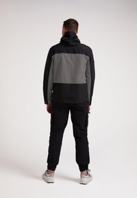 Giacca impermeabile nera e grigia con cappuccio, caratterizzata da un design a blocchi di colore e un tessuto testurizzato. Abbinata a pantaloni cargo neri.