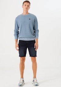 Suéter azul con canalé y cuello redondo, adornado con un parche de logo, combinado con pantalones cortos de mezclilla oscuros con el dobladillo vuelto. Calzado: zapatillas deportivas gris claro.