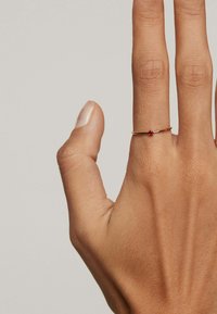Anillo de oro con una pequeña piedra roja, montado en una banda delgada, exhibido en una mano con textura de piel detallada y uña natural.