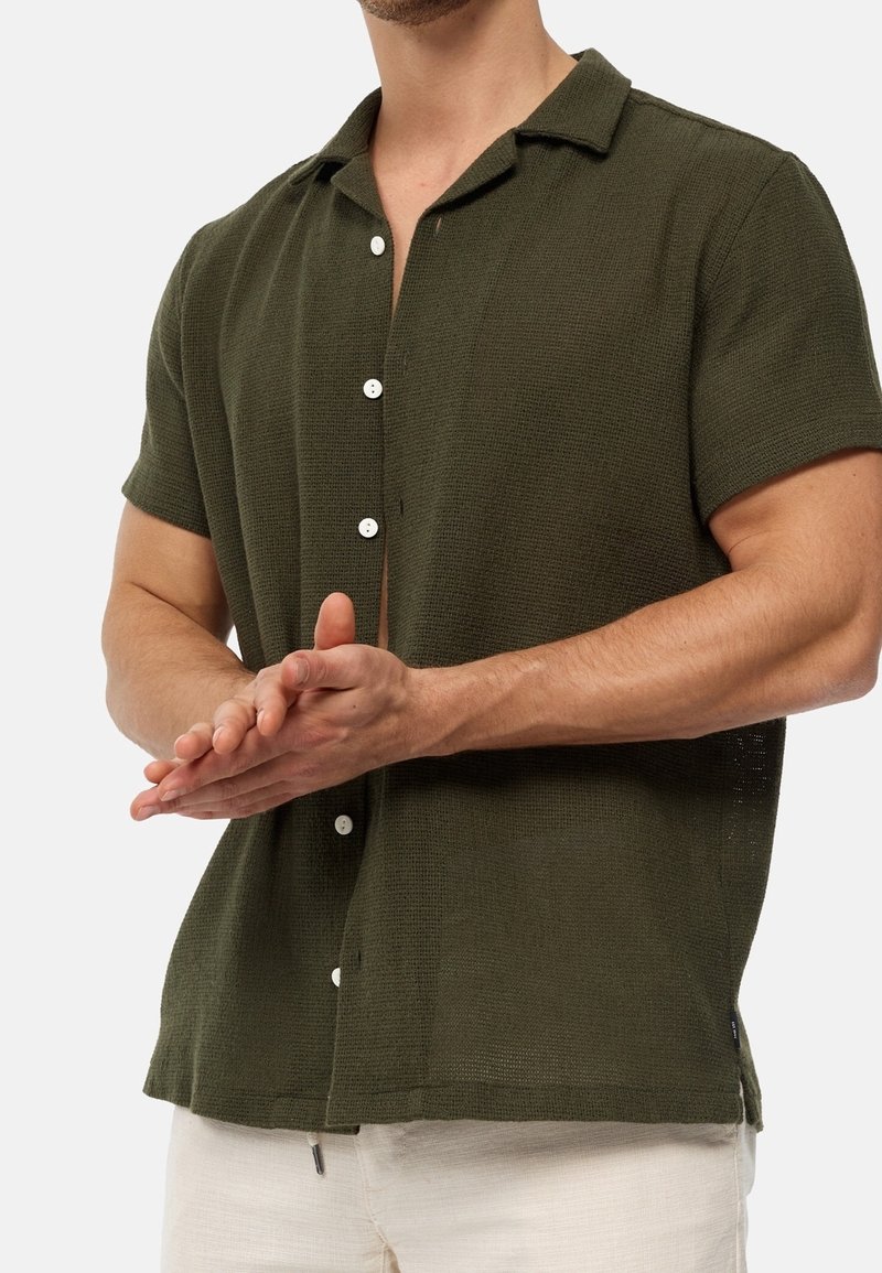 Chemise vert olive à manches courtes en tissu texturé, dotée d'un col classique et de boutons blancs, présentée avec les mains en geste d'applaudissement.