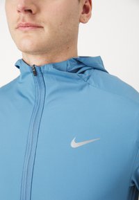 Lystblå Nike hættetrøje med fuld lynlås, blød tekstur og et reflekterende sølvlogo på venstre brystområde.