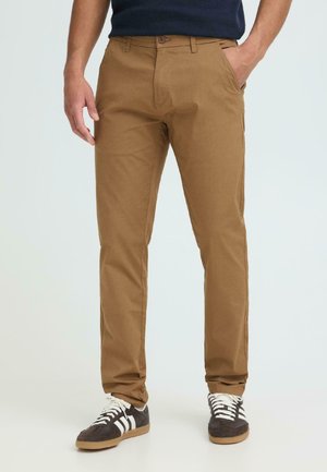 Mann trägt braune, eng anliegende Hosen mit Seitentaschen, schwarz-weiße Turnschuhe und ein dunkelblaues Hemd, steht vor einem schlichten Hintergrund.