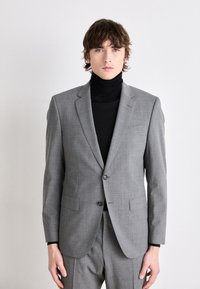 Blazer grigio con tessuto testurizzato, rever a intaglio, due tasche frontali e chiusura a due bottoni, indossato sopra un dolcevita nero.