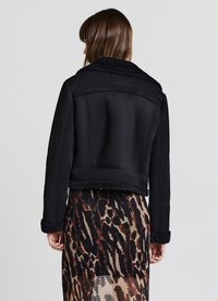 Giacca nera corta con colletto e polsini in tessuto testurizzato e peloso, caratterizzata da un tessuto liscio e cuciture strutturate, indossata sopra una gonna con motivo.