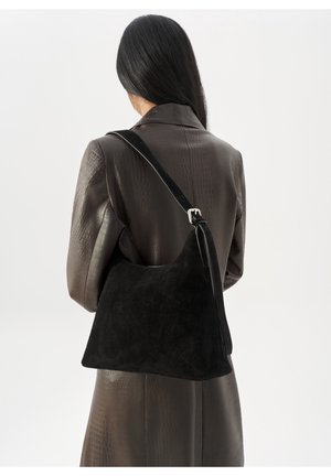 Kvinde med langt mørkt hår iført en tekstureret brun læderfrakke, der bærer en stor sort ruskind skuldertaske med et spænde.