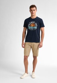 Tricou bleumarin cu mânecă scurtă, având un soare grafic și text, combinat cu pantaloni scurți bej. Tenisi albi completează look-ul casual.