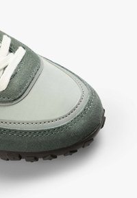 Grüner Wildleder- und synthetischer Laufschuh mit weißen Schnürsenkeln und einer strukturierten schwarzen Gummisohle, die ein schichtweise Materialdesign und Nähdetails präsentiert.