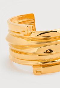 Βραχιόλι μανσετό σε χρυσό τόνο με στρωματοποιημένο, κυματιστό σχέδιο. Διαθέτει λείο, γυαλιστερό φινίρισμα και ασφαλές κούμπωμα στη μία άκρη.