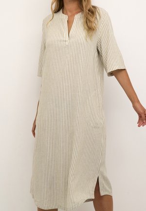 Kobieta ubrana w beżowo-białą sukienkę w pionowe paski o długości do kolan z rękawami do połowy ramienia i bocznym rozcięciem, stoi na tle gładkiego tła.