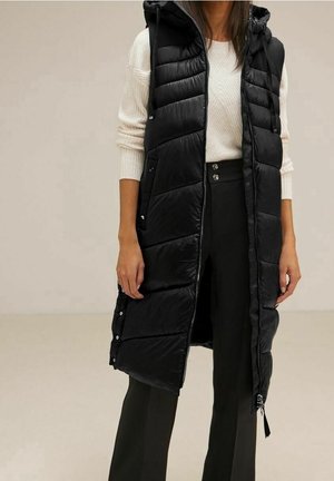 Vrouw draagt een mouwloze zwarte gewatteerde lange bodywarmer over een crèmekleurige trui en zwarte hoge taille wijde broek.