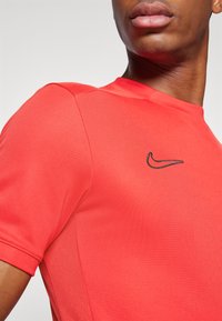 Rød treningsskjorte med rund hals, korte ermer og svart brodert Nike-logo på venstre bryst. Pustende nettpanel.