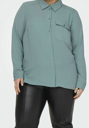 Persoon die een losse groene blouse met borstzak draagt en zwarte leren broek, staand tegen een eenvoudige lichte achtergrond.