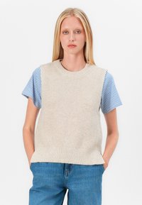 Haut sans manches en tricot beige avec un ourlet côtelé, superposé à une chemise à manches courtes bleu et blanche à rayures. Modèle portant un jean clair.