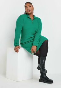 Vestido de punto verde con cuello con cremallera, que presenta mangas largas. Combinado con botas negras de caña alta y suela gruesa. Fondo blanco minimalista.