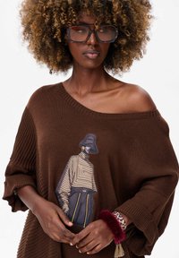 Bruine gebreide oversized trui met een off-shoulder ontwerp, voorzien van een grafische illustratie van een figuur. Accessoires omvatten een bont armband.
