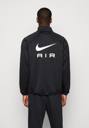 Personne portant une veste et un pantalon noirs, dos face à la caméra, avec un grand logo Nike Air blanc dans le dos de la veste.