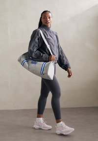 Mala de lona cinza com riscas azuis e logótipo "nike"; modelo em casaco cinza com fecho, leggings cinzas e ténis rosa claro. Texturas suaves.