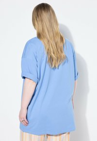 Svetle modré oversized tričko s ohrnutými rukávy, vyrobené z měkké tkaniny. Zadní strana má hladkou texturu bez vzorů nebo akcentů.