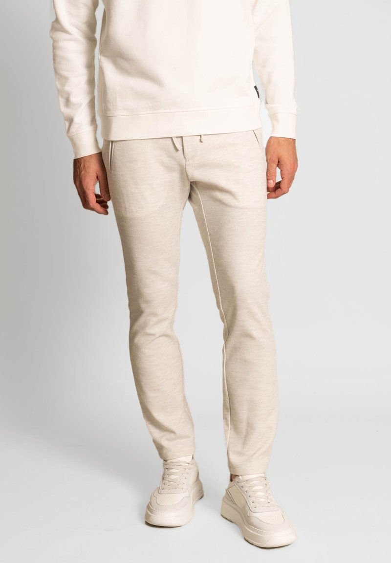 Béžové jogger kalhoty vyrobené z měkké tkaniny, s úzkým střihem, elastickým pasem a bočními kapsami, v kombinaci se světlými teniskami.