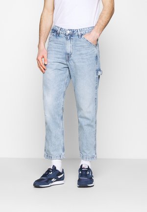 Mann trägt hellblaue Straight-Leg-Jeans, weiße Socken, marineblaue Sneakers und ein weißes T-Shirt, eine Hand in der Tasche.