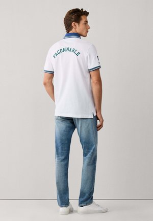 Homme portant un polo blanc avec un texte vert dans le dos et un col bleu, jean bleu clair et baskets blanches, debout devant un fond uni.
