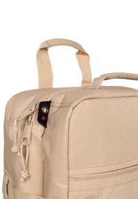 Beige tygryggsäck med fyrkantig form, utrustad med ett handtag högst upp, dragkedjestängning och ett svart metalldetalj på sidan.