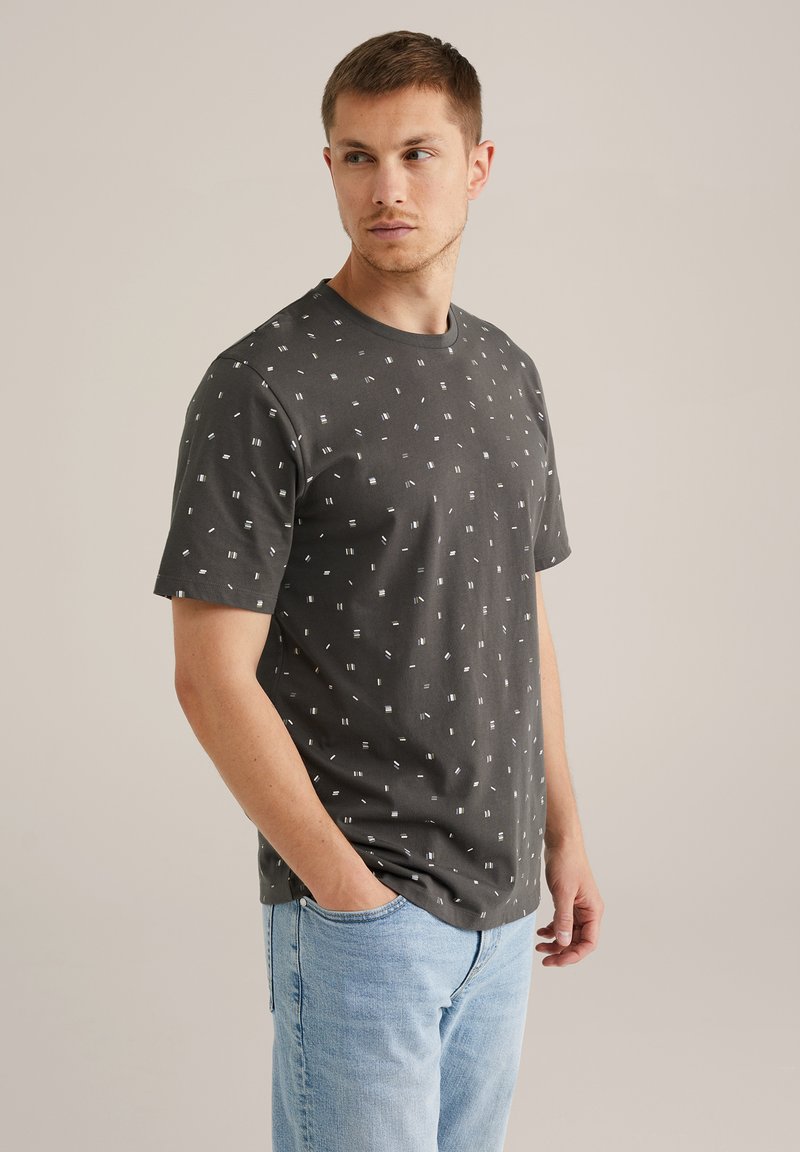 Grå T-shirt med korte ærmer lavet af bomuld med et spredt hvidt geometrisk mønster; rund halsudskæring; parret med lyseblå jeans.