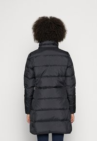 Osoba s kudrnatými vlasy, oblečená v dlouhé černé prošívané péřové bundě, zobrazená zezadu na jednobarevném světlém pozadí.