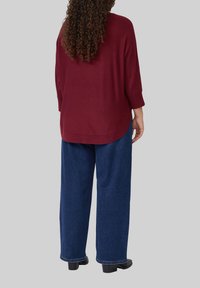 Burgunderfarbene, locker sitzende Bluse mit Dreiviertelärmeln, kombiniert mit hochgeschnittenen, dunkelblauen, weit geschnittenen Jeans. Weiche Textur, abgerundeter Saum.