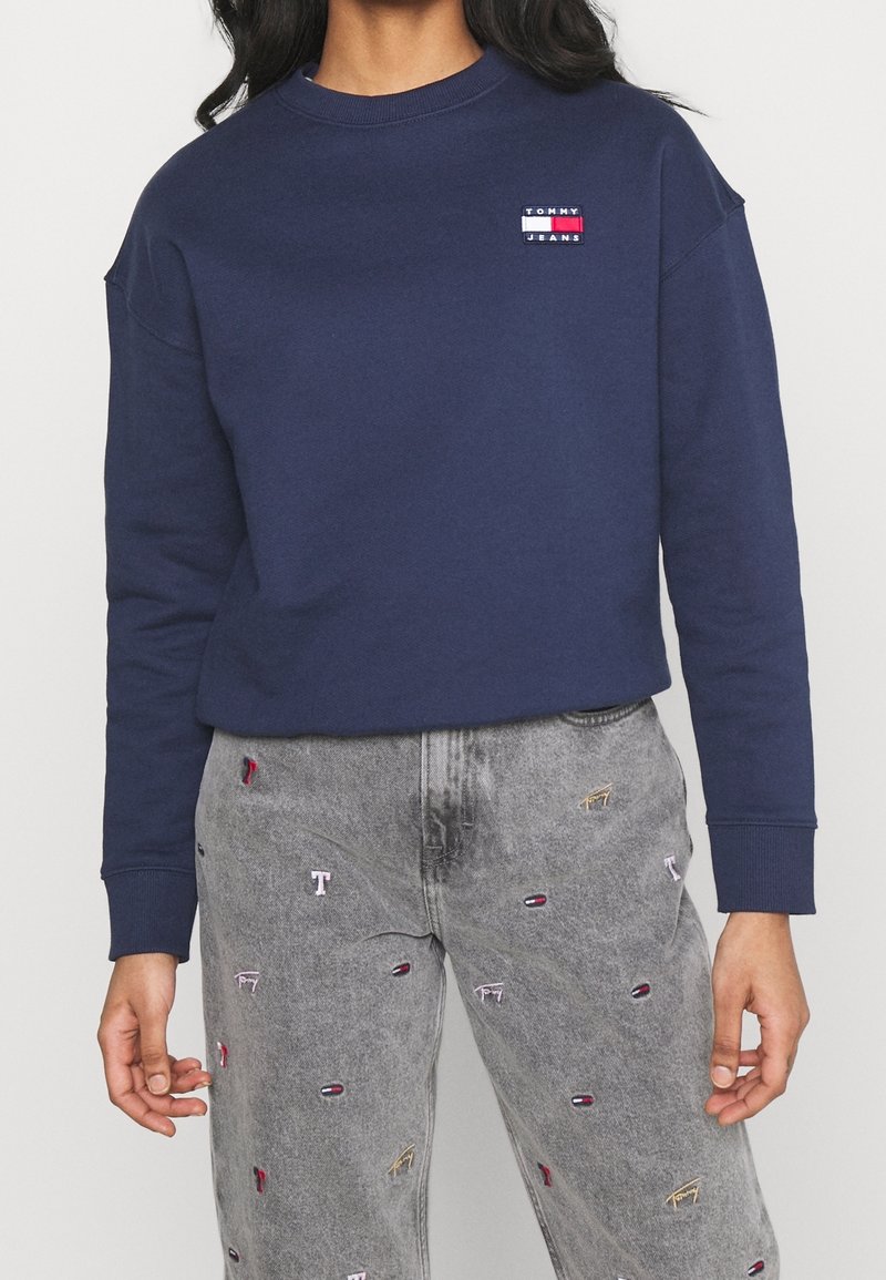 Marinblå sweatshirt med ribbad halsringning och broderad "TOMMY JEANS"-logotyp. Matchas med grå jeans med färgglada broderade detaljer.