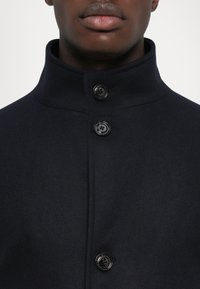 Palton din lână albastru marin cu guler înalt, având trei nasturi negri și o textură netedă. Prim-plan al zonei pieptului care arată detaliile nasturilor.