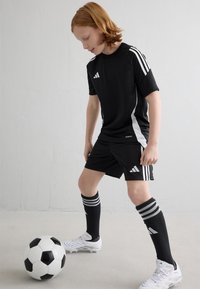 Equipación de fútbol negra de Adidas con rayas blancas, pantalones cortos negros, medias altas con rayas y botas blancas, colocada junto a un balón de fútbol.