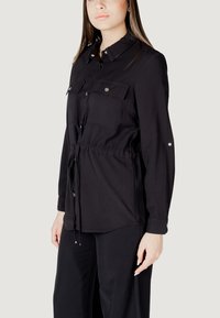 Camicia nera a maniche lunghe con colletto, vita regolabile con cordino, due taschini sul petto e dettagli in bottone argentato, realizzata in tessuto leggero.