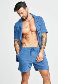 Camicia corta azzurra con texture, maniche corte, bottoni e pantaloni corti con coulisse abbinati. Presenta una vestibilità comoda e tasche laterali.