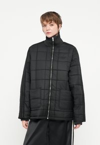 Casaco preto isolado com um padrão acolchoado, gola alta, fecho de zip, e dois bolsos frontais. Apresenta detalhes às riscas nas mangas.