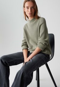 Giovane donna seduta su una sedia nera, che indossa un maglione lavorato a maglia verde chiaro e jeans neri a gamba larga, contro uno sfondo grigio uniforme.