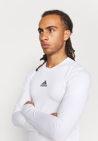 Weißes Langarm-Sportoberteil mit Rundhalsausschnitt und schwarzem Adidas-Logo in der Mitte; glatte Textur, taillierter Schnitt und keine sichtbaren Muster.